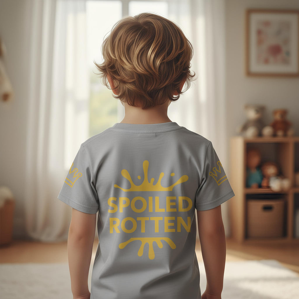 Child wearing a gray t-shirt with 'Spoiled Rotten' in gold text, standing in a room with toys and a window.