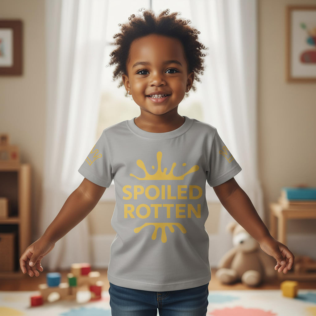 Child wearing a gray t-shirt with 'SPOILED ROTTEN' in gold, standing in a room with toys and a teddy bear.